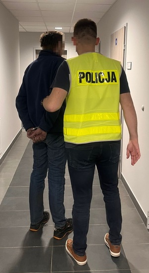 policjant i zatrzymany mężczyzna stojący tyłem do obiektywu aparatu, policjant ubrany w żółtą kamizelkę odblaskową z napisem policja