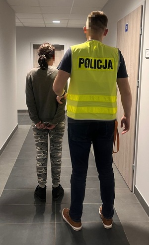 policjant i zatrzymana kobieta stojący tyłem do obiektywu aparatu, policjant ubrany w żółtą kamizelkę odblaskową z napisem policja
