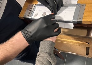 na zdjęciu widoczne dłonie policjanta ubrane w czarne rękawiczki jednorazowe, oraz dłoń kobiety, dodatkowo widoczny stolik do daktyloskopii, oraz odciski palców odbite na białej kartce papieru