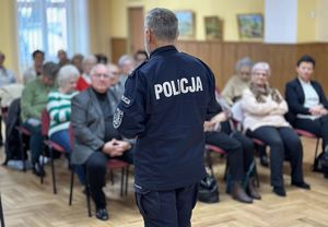 Sala lustrzana Miejskiego domu kultury w Oleśnie. Umundurowany policjant prowadzi pogadankę z seniorami. Słuchacze siedzą przed policjantem na krzesłach ustawionych w kilku szeregach.