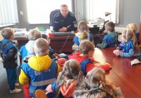 Policjant w mundurze siedzi za dużym biurkiem i opowiada dzieciom zgromadzonym wkoło o bezpieczeństwie.