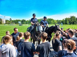 Zdjęcie przedstawia policjantów na koniach, przed nimi stoi zgromadzona młodzież.